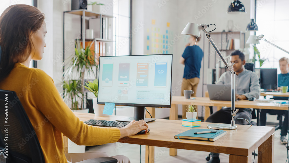 Poster Over the Shoulder: Creative Young Woman Sitting at Her Desk Using Desktop Computer with Screen Showing Project Management, Statistics and Graphs. Office with Diverse Team of Professionals Working.
