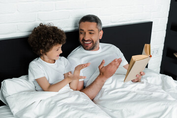 Obraz premium curly boy gesturing near happy father holding book in bedroom