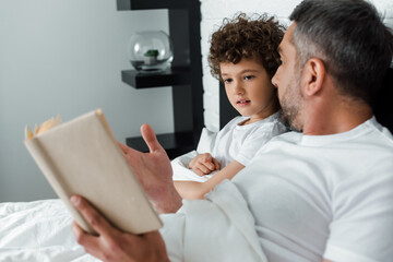 selective focus of curly boy looking at book near father in bedroom