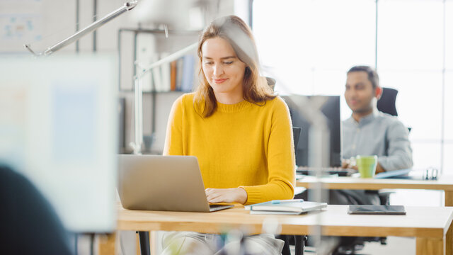 Beautiful and Creative Young Woman Sitting at Her Desk Using Laptop Computer. In the Background Bright Office where Diverse Team of Young Professionals Work.