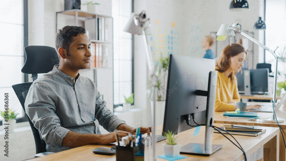 Canvas Prints Smart and Handsome Indian Office Worker Sitting at His Desk works on a Laptop. In the Background Modern Office with Diverse Team of Young Professionals Working.