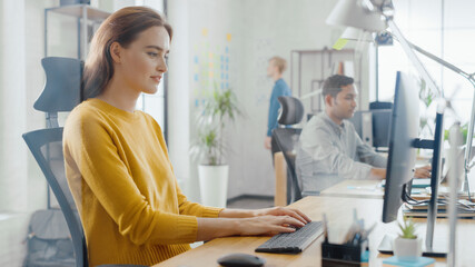 Beautiful and Creative Young Woman Sitting at Her Desk Using Laptop Computer. In the Background Bright Office where Diverse Team of Young Professionals Work.