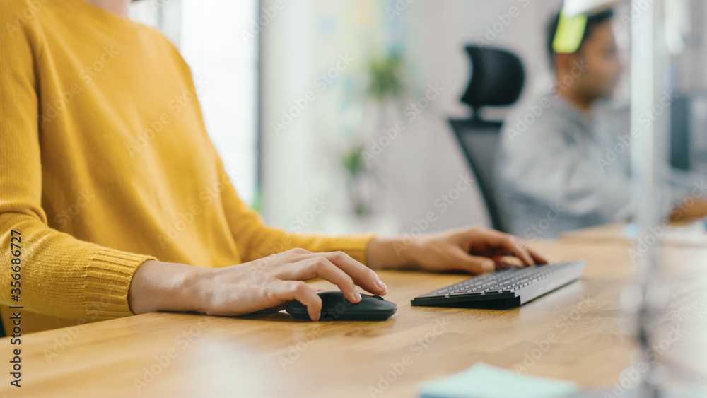 Poster Anonymous Young Woman Sitting at Her Desk Using Laptop Computer. Focus on Hands Using Mouse and Keyboard. In the Background Bright Office where Diverse Team of Young Professionals Work.