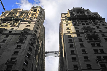 Haute passerelle en fer forgé et transparente reliant deux hauts bâtiments de style ancien vus en contre-plongée sur fond de ciel bleu et nuage blanc.