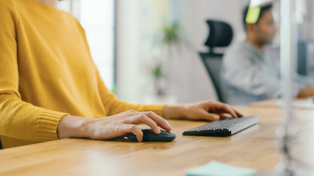 Anonymous Young Woman Sitting at Her Desk Using Laptop Computer. Focus on Hands Using Mouse and Keyboard. In the Background Bright Office where Diverse Team of Young Professionals Work.