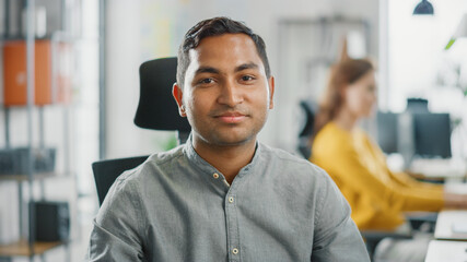Obraz premium Portrait of Handsome Professional Indian Man Working at His Desk, Using Personal Computer and Smiling at the Camera. Successful Man Working in Bright Diverse Office.