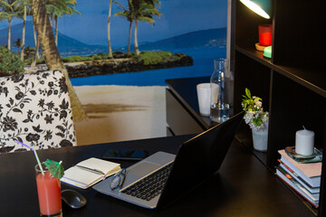 laptop with a mouse, a notebook and a glass of tropical cocktail with an umbrella on a black table. home work place in a room with bright blue photo wallpaper with palm trees and the sea.