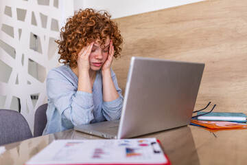 Young frustrated woman working at office desk in front of laptop suffering from chronic daily...