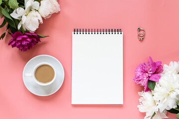 Stylized female office desktop with blank notepad, jewelry set, cup of coffee and peony flowers on pink background top view.