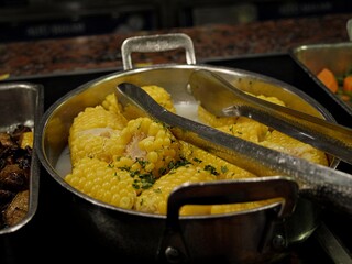 Boiled corn on the cob set at a buffet with a pair of silver tongs at a restaurant
