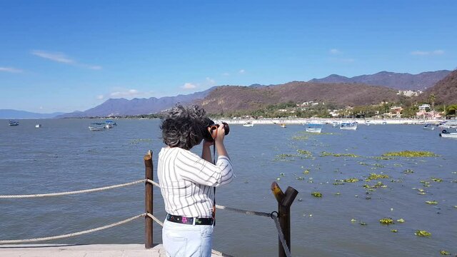 Female tourist taking a picture of Lake Chapala with calm waves, lily, boats and with the boardwalk and village in the background, sunny day in Jalisco, Mexico