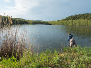 Obraz na płótnie Canvas Fly Fisherman angler standing on shore of calm water of forest lake, fish pond Kunraticky rybnik with birch and spruce trees growing along the shore. Nature fishing background. Springtime