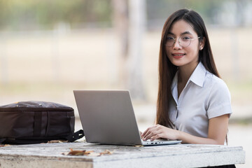 Young teen female Asian university student sits on bench typing laptop in the university campus. Female university student go to university. Back to school concept.