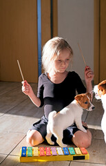 girl plays a melody on a xylophone sitting on her knees in a gymnastic suit with a puppy of Jack Russell Terrier and an adult dog in the room. self-isolation, vertical format