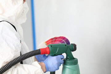 Close-up of professional worker painting wall with spray gun. Person in white protective costume and blue gloves. Renovation and construction site concept