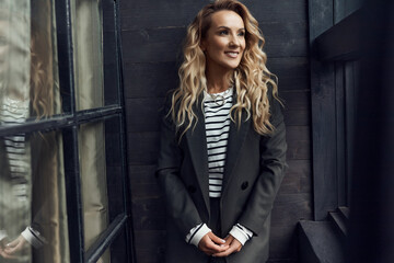 Fototapeta na wymiar photo of a beautiful woman with long blond curly hair and professional makeup standing, looking away and smiling, she is wearing a black suit and striped blouse