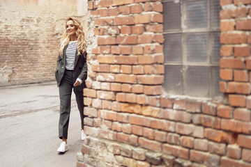 streetstyle photo of a beautiful woman with long blond curly hair who comes out of the corner and smiles, she is wearing a black suit, striped blouse and white sneakers, her hand in her pocket