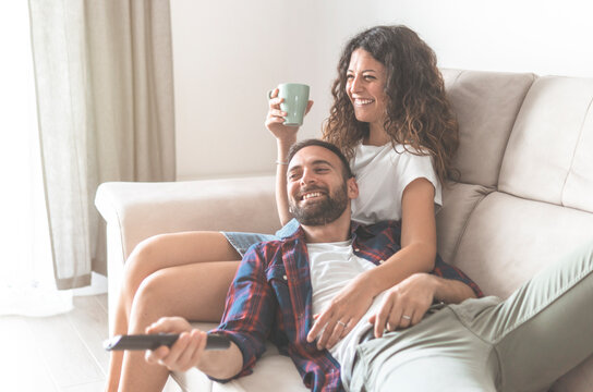 Young couple sitting on the sofa watching tv and drinking a cup of coffee - lovers relax and enjoy free time at home.