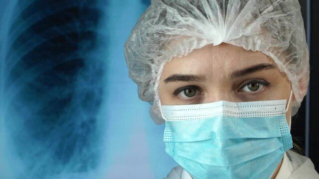 Portrait of medic woman on background of X-ray of lungs with pneumonia. Nurse looks at camera and corrects medical mask hands in medical gloves. Close-up. Coronavirus. COVID-19.