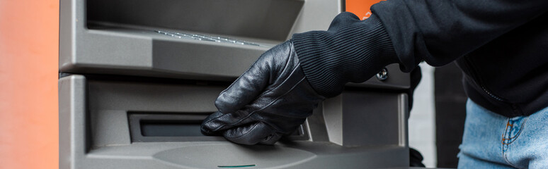 Panoramic crop of burglar holding hand near cash dispenser of atm