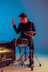 Caucasian male drummer improvising isolated on blue studio background in neon light. Performing, looks inspired, energy. Concept of human emotions, facial expression, ad, music, art, festival.