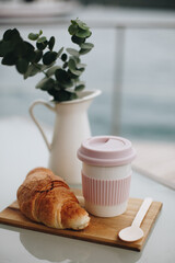 Cup of coffee on wooden background with croissant and white vase with greens. Breakfast or coffee brake theme.