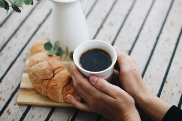 Cup of coffee on wooden background with croissant and white vase with greens. Breakfast or coffee brake theme.
