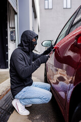 Side view of robber in balaclava using screwdriver while opening lock on door of car on urban street
