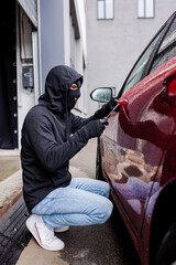 Obraz na płótnie Canvas Side view of robber in balaclava using screwdriver while opening lock on door of car on urban street