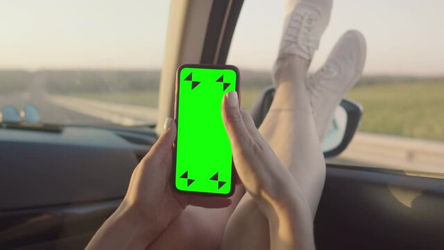 The girl in the front seat of the car is sitting with a green screen phone with her feet out the window