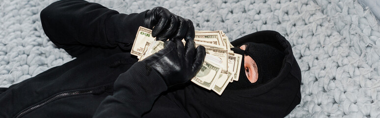 Panoramic shot of thief in balaclava holding money near face on bed