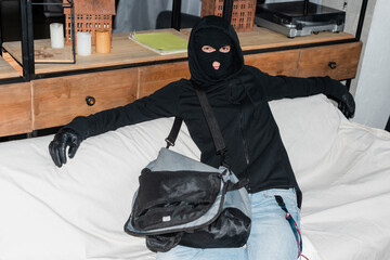 Robber in balaclava looking at camera while sitting near laptop in bag