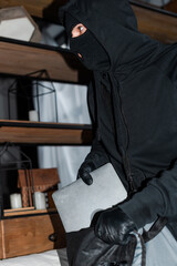 Selective focus of robber in mask and leather gloves putting laptop in bag