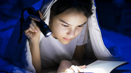 Portrait of girl lying under blanket at night and reading book with light torch