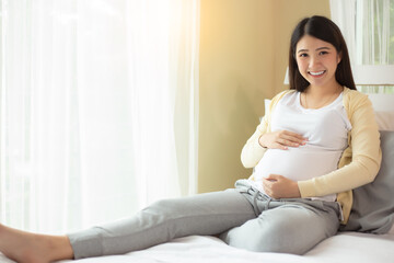 Happy pregnant young asian woman show big belly, admiring her baby or fetus on bed with happiness smile face. Young Asia mother love her baby so much. Mom expecting baby has good health smiley face