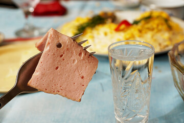      Sausage strung on a fork next to a wineglass of vodka on the background of a table with snacks and dishes.