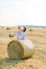 Happy two year old girl child in striped dress playing with young smiling mom in summer autumn field with hay bales, sitting together on hay stack. Summer sunset portrait