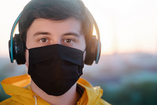 A young man in a medical mask black , a jacket bright yellow, and large headphones looks ahead against the blurry background of the city