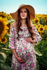 A young pregnant brunette woman in a straw hat and a light dress, walks along the field of sunflowers in the summer. The concept of a happy expectation of motherhood, the girl is happy and smiling.