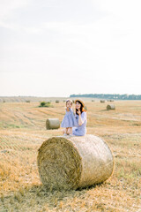 Naklejka na ściany i meble Happy two year old girl child in striped dress playing with young smiling mom in summer autumn field with hay bales, sitting together on hay stack. Summer sunset portrait