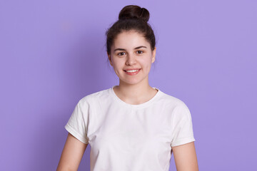 Sensual young woman with hair bun against lilac background. Attractive female model with perfect skin and charming smile, wearing white casual t shirt, teenage with positive emotions.