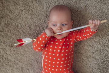 cupid and arrow, сute baby in a red bodysuit with white hearts lies on the carpet and holds an...
