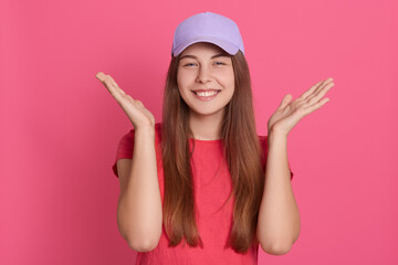 Obraz na płótnie Canvas Close up portrait of beautiful young caucasian woman poses smiling indoors, spreading hands aside, wearing casual t shirt and baseball cap,standing against pink wall.
