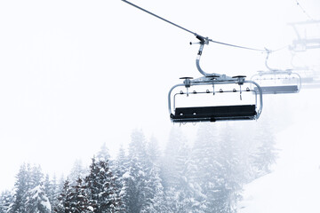 An empty chairlift appearing through the foggy cloud layer on a mountainside. Snow covered trees...