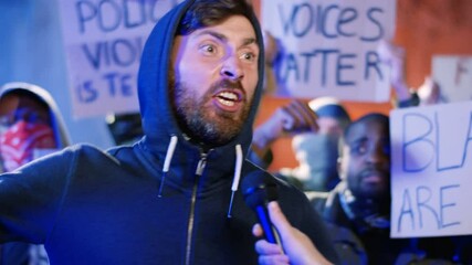 Caucasian female correspondent interviewing aggressive male protester at street at night while people protesting for human rights and against police violence. Interview of rebel man by woman for news.