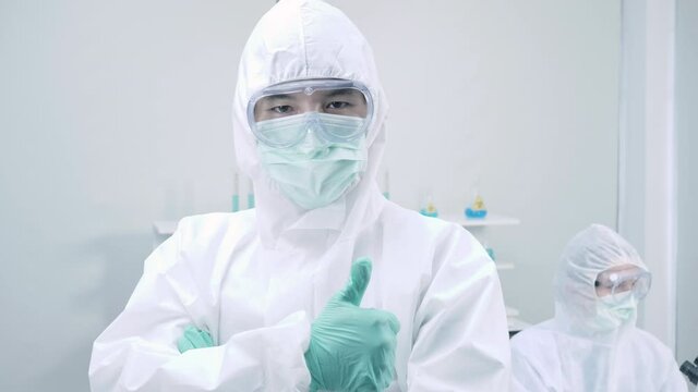 Asian man scientist is wearing Personal Protective Equipment (PPE) looking at camera while standing with his arms crossed and Showing thumb up in a laboratory
