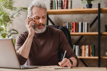 Handsome businessman in casual clothes talking on mobile phone and making notes while sitting in his office - Powered by Adobe