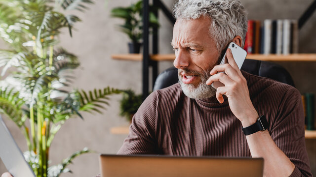 Annoyed senior man argue over phone explaining his point of view to another person while sitting at workplace