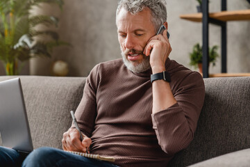 Middle aged businessman in casual talking on mobile phone and writting notes while working at home