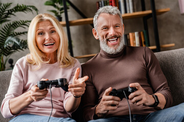 Laughing middle aged couple playing video games with joystick sitting on couch in living room